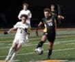 Two high school soccer players in mid-action during a night game, one in white, the other in black and gold, on a field.
