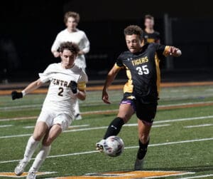 Two high school soccer players in mid-action during a night game, one in white, the other in black and gold, on a field.