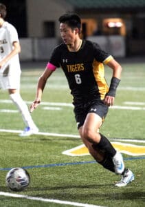 Soccer player in black Tigers jersey dribbling ball on field during night match.