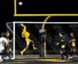 Soccer goalie in yellow leaps to save a goal during a night match, teammates watching intently.