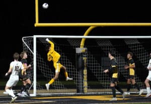 Soccer goalie in yellow leaps to save a goal during a night match, teammates watching intently.
