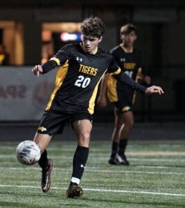 Soccer player in a black and yellow Tigers uniform kicks a ball during a night game on the field.