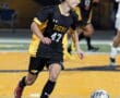 Soccer player in black and yellow jersey dribbles the ball during a night game on a turf field.