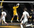 Goalkeeper jumps to catch soccer ball during night match, players in white and black watching.