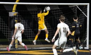 Goalkeeper jumps to catch soccer ball during night match, players in white and black watching.