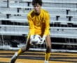 Goalkeeper in yellow kit catching soccer ball during a game, with stadium seats in the background.