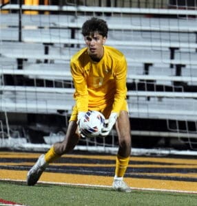 Goalkeeper in yellow kit catching soccer ball during a game, with stadium seats in the background.