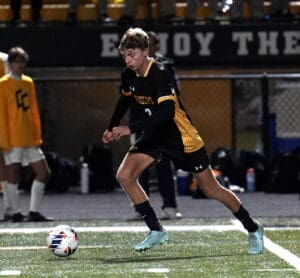 Soccer player in black and yellow uniform dribbling the ball under stadium lights during a match.
