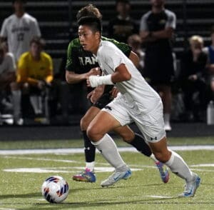 Soccer player in white kit dribbling ball during intense match, with opponent behind, on a green field at night.