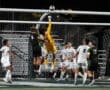 Soccer match action at goalpost with goalkeeper leaping, players watching, under stadium lights.