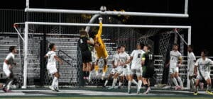 Soccer match action at goalpost with goalkeeper leaping, players watching, under stadium lights.
