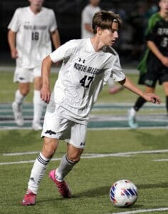 Soccer player in white North Allegheny uniform dribbling a ball on the field during a game.