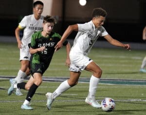 Boys high school soccer match, one player dribbling the ball while others are in pursuit on a grassy field.