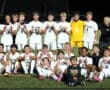 Youth soccer team celebrates on the field, smiling in white uniforms with a coach and child in front.
