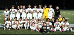 Youth soccer team celebrates on the field, smiling in white uniforms with a coach and child in front.