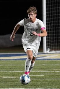 Soccer player in white uniform dribbles ball on field at night, wearing pink armband and focused expression.