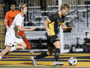 Two soccer players in action during a night game, one in white chasing another in black and yellow near the goal.