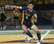 Soccer player in black and gold uniform dribbles ball on the field during a night game.
