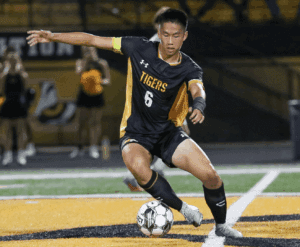 Soccer player in black and gold uniform dribbles ball on the field during a night game.