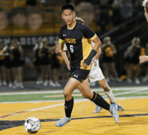 Soccer player dribbling the ball during a game on a field at night in a black and yellow uniform.