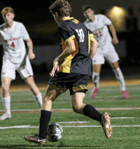 Soccer player in black and yellow dribbling ball during night match, opponents in white nearby.