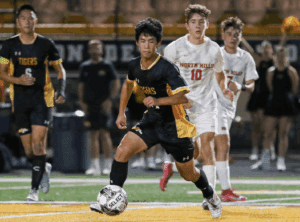 Soccer player dribbles ball during a high school match, with teammates and opponents in pursuit on the field.
