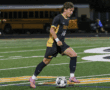 Soccer player in black and yellow uniform dribbles ball on field during night game.