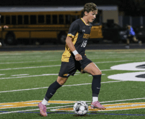 Soccer player in black and yellow uniform dribbles ball on field during night game.