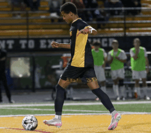 Soccer player in black and yellow uniform dribbles ball on field during nighttime match, with team bench in background.