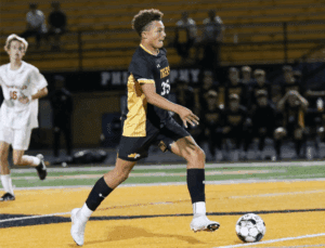 Soccer player in black and yellow uniform dribbles ball on field during a match.