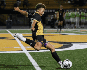 Soccer player in striped jersey kicking ball on field during match, with blurred players in the background.