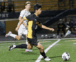 Soccer player in black and yellow uniform dribbles ball during night match on green field.