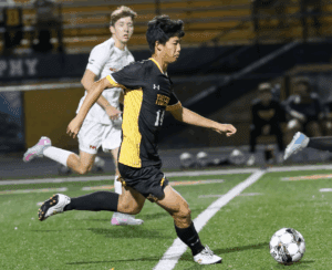 Soccer player in black and yellow uniform dribbles ball during night match on green field.