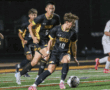 High school soccer game action with players in black and yellow uniforms dribbling on the field.
