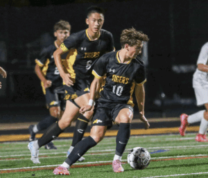High school soccer game action with players in black and yellow uniforms dribbling on the field.