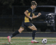Soccer player in black and yellow uniform dribbles ball on field during night match.