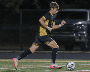 Soccer player in black and yellow uniform dribbles ball on field during night match.