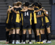 Soccer team huddles on field in black and yellow uniforms, showing unity and team spirit.