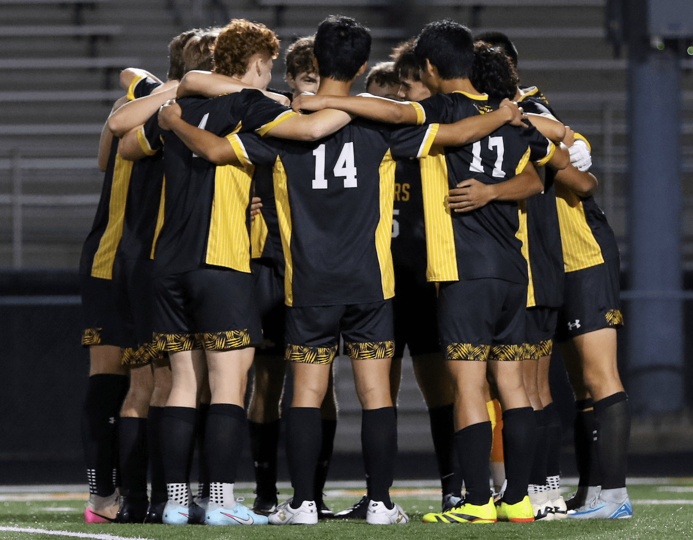 Soccer team huddles on field in black and yellow uniforms, showing unity and team spirit.