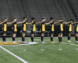 Soccer team lining up on the field at night, wearing black and yellow uniforms with a stadium in the background.