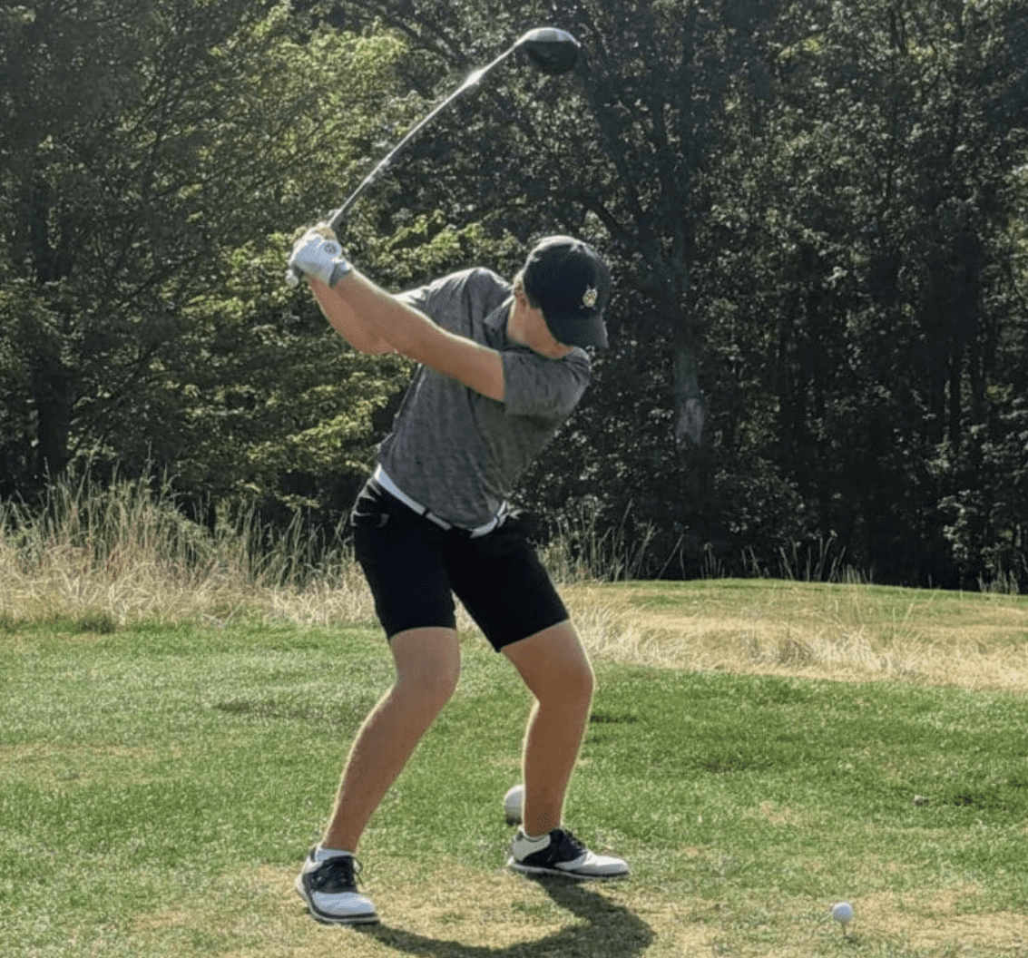 Golfer in mid-swing on a sunny course, surrounded by green trees and grass.