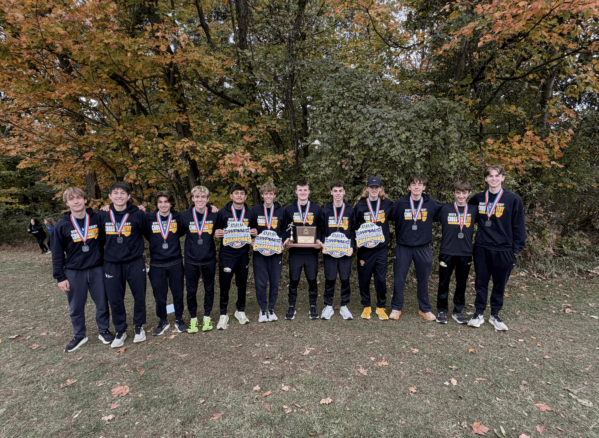 High school cross country team celebrates championship with medals and a trophy, standing in front of autumn trees.