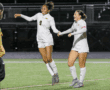 Two female soccer players in white uniforms celebrating on the field with smiles and holding hands.