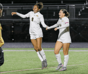 Two female soccer players in white uniforms celebrating on the field with smiles and holding hands.