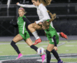 Soccer player in white jersey leaps over two players in green during a match on a stadium field.
