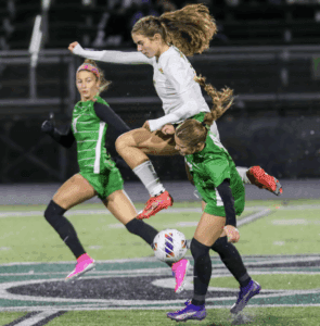 Soccer player in white jersey leaps over two players in green during a match on a stadium field.