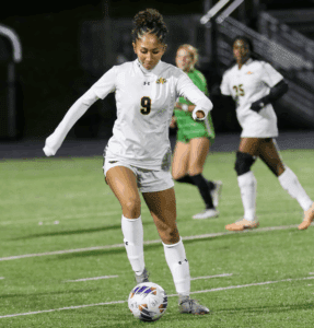 Soccer player in white jersey dribbling on the field during a night match, with teammates in the background.
