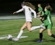 Soccer player in white jersey kicking ball during intense match on green field at night.