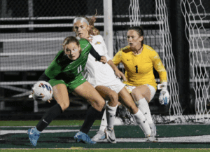 Soccer players compete intensely near goalpost during match, focused on ball control and defense strategy.