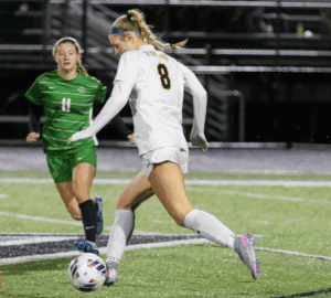 Soccer player in white jersey dribbling ball, opponent in green approaching on field.